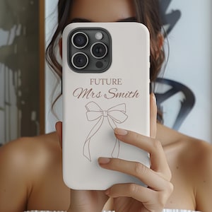 May include: A cream-coloured iPhone case featuring the text "FUTURE Mrs Smith" and a bow illustration in brown. The case has a camera cutout and is held in front of a person.