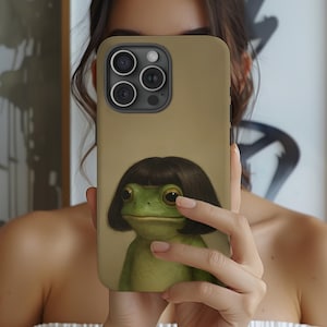 May include: A phone case with a tan background featuring a green frog with a dark brown bob haircut. The frog has large, expressive eyes and is centered on the case. The phone case has multiple camera lenses.