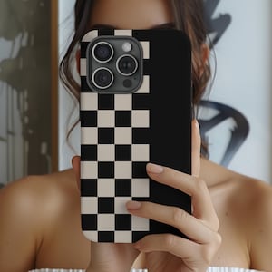 May include: A black and cream checkered phone case. The case has a black top half with the camera lenses visible. The bottom half features a checkerboard pattern in black and cream. The phone case is held in front of a person's face.