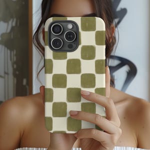 May include: A smartphone case with a cream-colored background and a pattern of olive green squares. The case has a camera cutout and is being held up in front of a person's face.