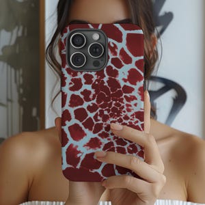 May include: A maroon and light blue phone case with a giraffe print design. The case has a dark camera module and is held in front of a person's face. The case has a smooth, glossy finish.