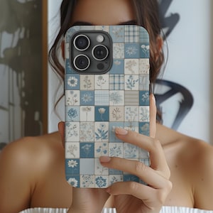 May include: A phone case with a floral tile design in shades of blue, white, and beige. The case features a grid pattern of squares, each displaying a different flower or plaid design. The phone case is held up in front of a person.
