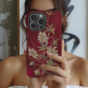 May include: Burgundy phone case with a floral design in white and green. The case has a textured finish and a dark camera module. The floral pattern features detailed flowers and leaves, creating an elegant aesthetic.