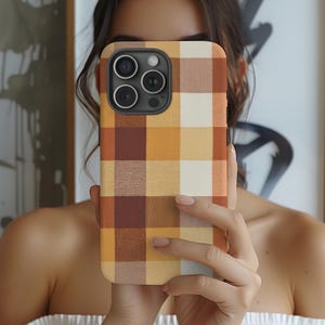 May include: A smartphone case featuring a classic plaid pattern in shades of warm brown, golden yellow, and cream. The design covers the entire back of the case, which includes a prominent black camera module with three circular lenses and a flash.