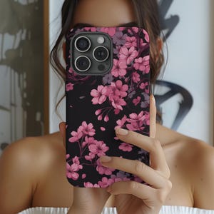 May include: Black smartphone case featuring a floral design with pink cherry blossoms. The case has a matte finish and a raised edge around the camera. The flowers are detailed with varying shades of pink and purple.