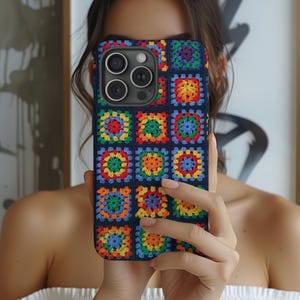 May include: A smartphone case featuring a colorful crochet granny square pattern. The design consists of multiple small, square motifs in vibrant shades of red, orange, yellow, green, blue, and purple, all bordered by a dark navy blue yarn.