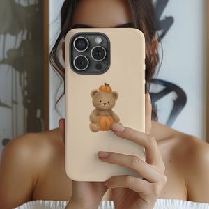 Könnte beinhalten: Eine beige Smartphone-Hülle mit einer zentralen Illustration eines braunen Teddybären, der mit einem kleinen orangefarbenen Kürbis auf dem Kopf und einem weiteren Kürbis zwischen den Beinen sitzt. Die Hülle hat ein schwarzes Kameramodul.