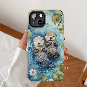 Puede incluir: Una funda para teléfono con un diseño de acuarela de dos nutrias nadando en una escena de agua azul y verde. Las nutrias son marrones y blancas y tienen caras lindas. La funda tiene un recorte para la cámara y un diseño de flores blancas.