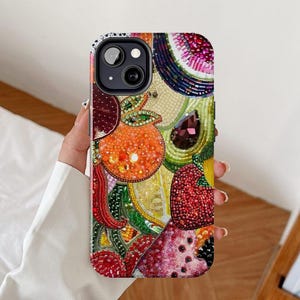 Pode incluir: Uma capa de telefone colorida com um design de frutas com contas. A capa apresenta um arranjo vibrante de frutas, incluindo laranjas, morangos e melancia, com intrincados trabalhos de contas em várias cores. A capa tem uma lente de câmera preta.