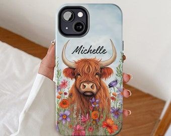 May include: A phone case with a watercolor design of a Highland cow with brown fur and a white face, standing in a field of colorful flowers. The name "Michelle" is written in black cursive above the cow.