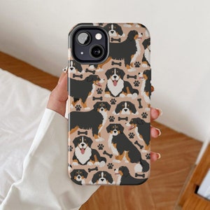 May include: A phone case featuring a pattern of Bernese Mountain Dogs on a light brown background. The case has a black camera lens cutout and a white outline around the dogs.