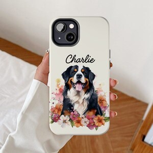 Può includere: Custodia bianca per iPhone con un design ad acquerello di un cane da montagna bernese con fiori. Il cane guarda la fotocamera e ha un'espressione felice. Il testo "Charlie" è stampato sulla custodia.