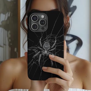 May include: Black phone case featuring a detailed spider web design with a spider in the center. The spider and web are in shades of gray against a black background. The case has a glossy finish and is designed for a smartphone.