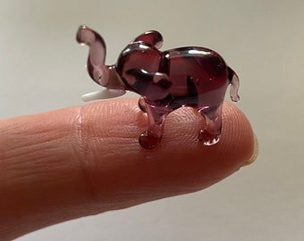 May include: A small glass elephant figurine in a deep purple color. The elephant has a white glass trunk and is standing on a finger.
