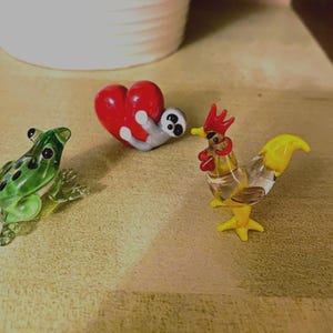 May include: A selection of glass ornaments: a green frog with black spots, a red heart with a grey creature, and a yellow and clear rooster. The figurines are displayed on a textured surface.