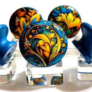 May include: A collection of decorative cabinet knobs. The knobs feature a vibrant floral design in shades of blue, yellow, and orange. Two blue, wing-shaped knobs flank three round knobs, all displayed on clear acrylic stands.