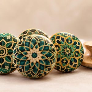 May include: A collection of round cabinet knobs featuring a circular mandala-style pattern in shades of teal, dark green, and gold. The knobs have a smooth, light-colored wooden base and a flat, decorative front surface with intricate geometric designs.