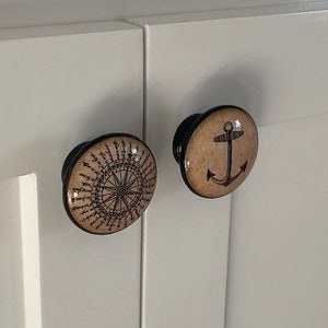 以下が含まれることがあります： 航海をテーマにした装飾的なキャビネットノブが2つ。1つのノブは、ベージュの背景に黒と白のデザインのコンパスローズが描かれています。もう1つのノブは、ベージュの背景に黒いアンカーが描かれています。