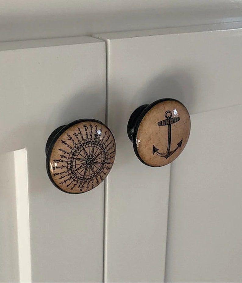 以下が含まれることがあります： 航海をテーマにした装飾的なキャビネットノブが2つ。1つのノブは、ベージュの背景に黒と白のデザインのコンパスローズが描かれています。もう1つのノブは、ベージュの背景に黒いアンカーが描かれています。