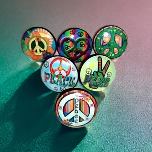 May include: A collection of six circular buttons featuring colorful peace sign designs. The buttons display various patterns including floral motifs, rainbow stripes, and the word PEACE in stylized lettering, all set against a glossy finish.