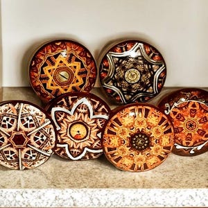 May include: A collection of seven decorative cabinet knobs with intricate, geometric designs. The knobs feature a mix of warm colors, including brown, gold, and orange, with detailed patterns. They are arranged on a light-colored surface.
