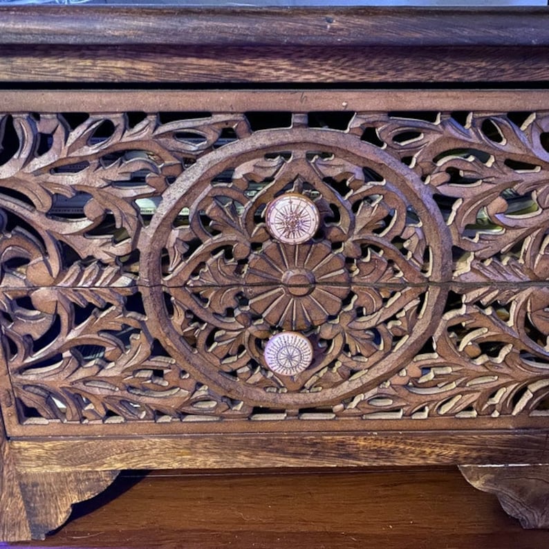 以下が含まれることがあります： 複雑な彫刻と2つの白いノブが付いた木製の箱。箱は濃い茶色の仕上げで、装飾的なデザインが施されています。