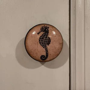 以下が含まれることがあります： 黒と白のタツノオトシゴのデザインが施された、クリーム色の丸いノブ。ノブはセラミック製で、光沢のある仕上げです。