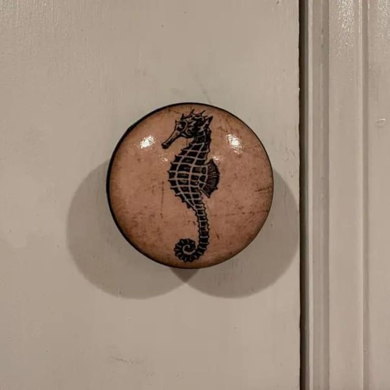 以下が含まれることがあります： 黒と白のタツノオトシゴのデザインが施された、クリーム色の丸いノブ。ノブはセラミック製で、光沢のある仕上げです。