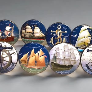 May include: A set of eight round cabinet knobs featuring nautical themes. The designs include various sailing ships, anchors, a ship's wheel, and a life preserver. Each knob has a glossy finish with a background of blue or white script text.