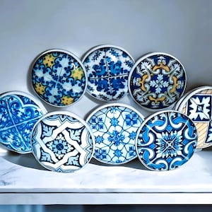 May include: A collection of eight round decorative cabinet knobs with intricate blue and white tile patterns. Each knob features a unique design with floral and geometric motifs, set against a white background. The knobs are arranged on a white surface.