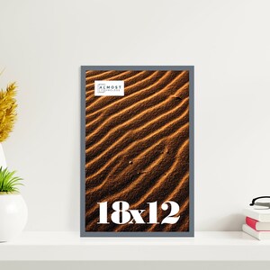 May include: A framed print featuring a close-up of rippled sand dunes in shades of brown and tan. The frame is a dark gray. The print is labeled "18x12" in white text. The top right corner of the frame has the text "ALMOST FRAMELESS".