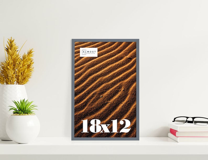 May include: A framed print featuring a close-up of rippled sand dunes in shades of brown and tan. The frame is a dark gray. The print is labeled "18x12" in white text. The top right corner of the frame has the text "ALMOST FRAMELESS".