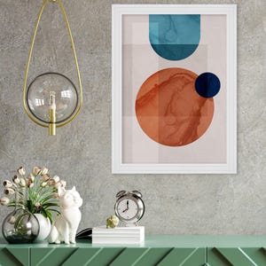 May include: A white framed print with an abstract design featuring a large orange circle with a smaller blue circle inside, and a blue curved shape above. The print is hanging on a gray wall above a green dresser with a white cat figurine and a silver alarm clock.