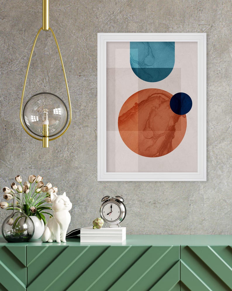 May include: A white framed print with an abstract design featuring a large orange circle with a smaller blue circle inside, and a blue curved shape above. The print is hanging on a gray wall above a green dresser with a white cat figurine and a silver alarm clock.