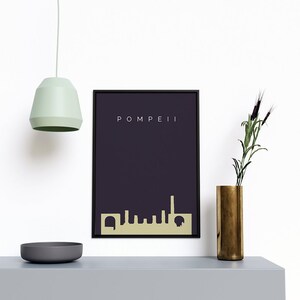 May include: A framed art print featuring the word "POMPEII" above a gold-colored skyline silhouette against a dark purple background. The print is in a black frame. A gold vase with greenery and a gray bowl are on a gray surface.
