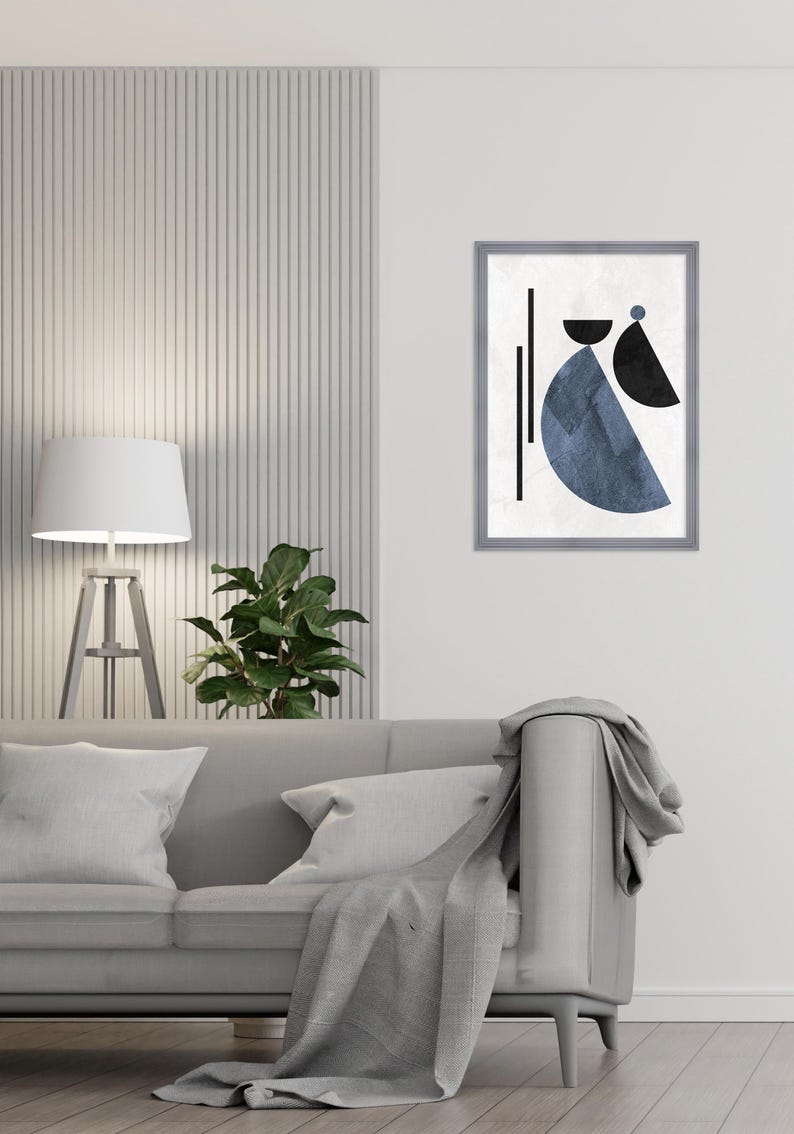 May include: A gray sofa with a throw blanket and two pillows. A white lamp with a white shade is on a wooden stand to the left of the sofa. A green plant is in a pot on the table. A framed print with a blue and black abstract design is on the wall behind the sofa.