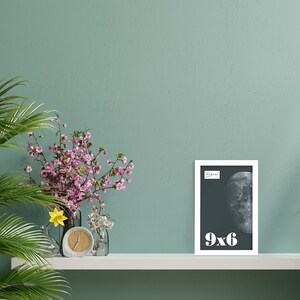 May include: A white framed 9x6 inch print featuring a grayscale image of the moon. The print is displayed on a white shelf against a teal wall. Decorative flowers and a clock are also on the shelf.
