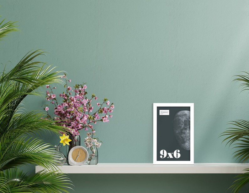 May include: A white framed 9x6 inch print featuring a grayscale image of the moon. The print is displayed on a white shelf against a teal wall. Decorative flowers and a clock are also on the shelf.