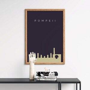 May include: A framed art print featuring the word "POMPEII" above a gold-colored skyline silhouette against a dark blue background. The print is in a wooden frame. A wooden hand sculpture and books are on a black table.