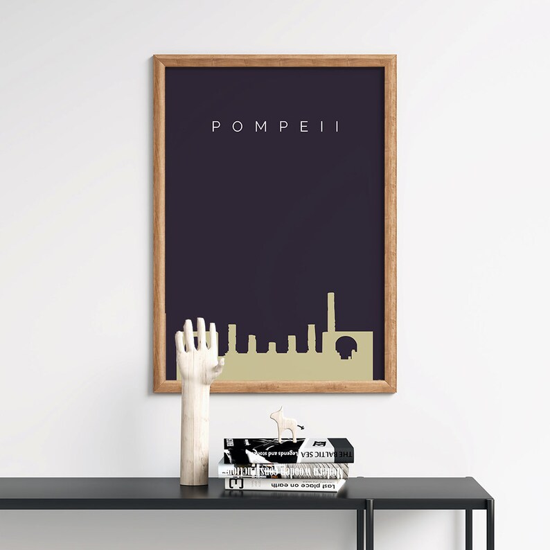 May include: A framed art print featuring the word "POMPEII" above a gold-colored skyline silhouette against a dark blue background. The print is in a wooden frame. A wooden hand sculpture and books are on a black table.