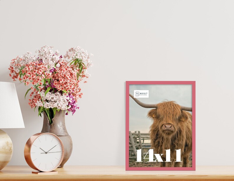 May include: A framed print featuring a Highland cow with long horns, set against a rural backdrop. The frame is a vibrant pink, and the print is labeled "14x11". A bouquet of flowers and a clock are also on the table.