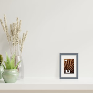 May include: A gray picture frame with a white mat and a brown and gold abstract image inside. The frame is sitting on a white shelf with a cactus, a succulent, and dried flowers in vases.
