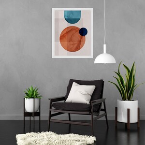 May include: A white framed print with an abstract design featuring geometric shapes in shades of blue, red, and white. The print is hanging on a gray wall above a black armchair with a white pillow. A white hanging lamp is above the chair and a potted plant is on the right side of the chair.