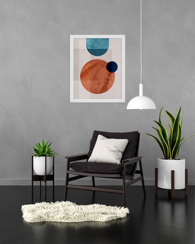 May include: A white framed print with an abstract design featuring geometric shapes in shades of blue, red, and white. The print is hanging on a gray wall above a black armchair with a white pillow. A white hanging lamp is above the chair and a potted plant is on the right side of the chair.