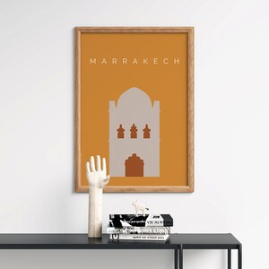 May include: A framed print with a minimalist design of a building in a muted orange color with the word "MARRAKECH" in white text at the top. The print is sitting on a black table with a wooden hand sculpture and a few books.