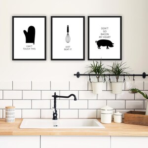 May include: Three black and white framed prints with kitchen-themed illustrations and text. The first print shows a black oven mitt with the text "Can't Touch This". The second print shows a black whisk with the text "Just Beat It". The third print shows a black pig with the text "Don't Go Bacon My Heart".