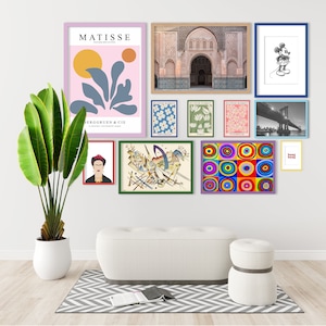 May include: A gallery wall featuring a selection of framed artwork, including a pink and blue abstract print, a black and white illustration of Mickey Mouse, a photograph of a bridge, and a colourful geometric print. The wall is decorated with a white sofa and a white stool.