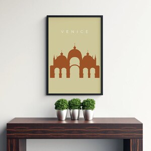 May include: A minimalist poster with a beige background and a black frame. The poster features a brown silhouette of a building with multiple domes, and the word "VENICE" in white text at the top.