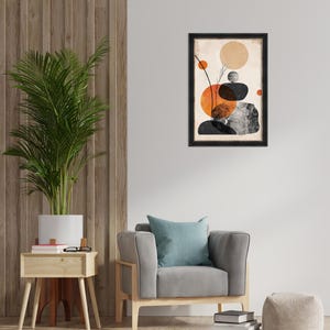 May include: Abstract art print in a black frame. The artwork features orange, black, and gray shapes on a beige background. The print hangs above a gray armchair with a blue pillow. A potted plant and side table are also in the room.