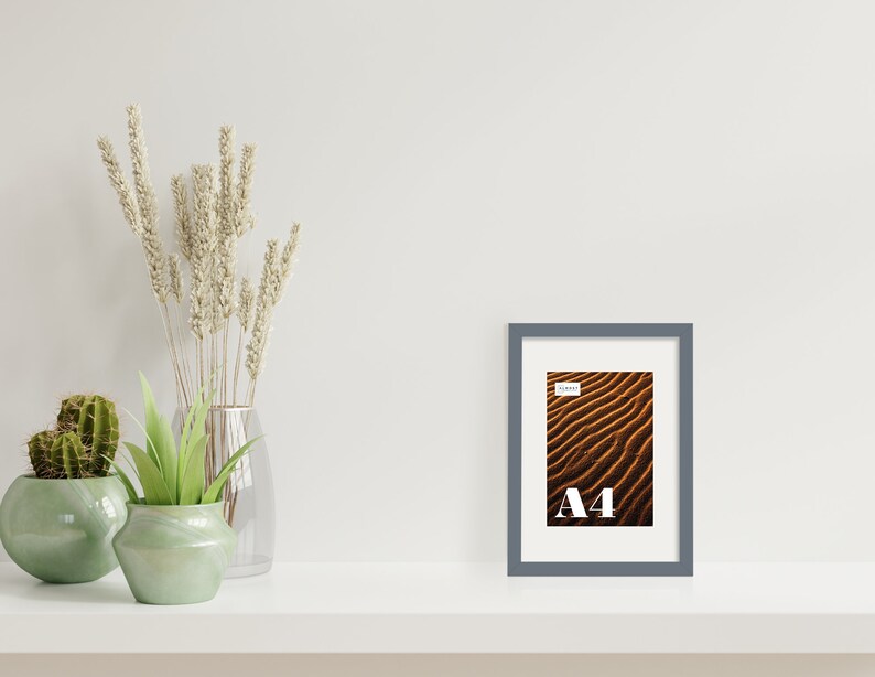 May include: A gray picture frame with a white mat and a photo of sand dunes. The frame is sitting on a white shelf with a vase of dried flowers and two potted plants.